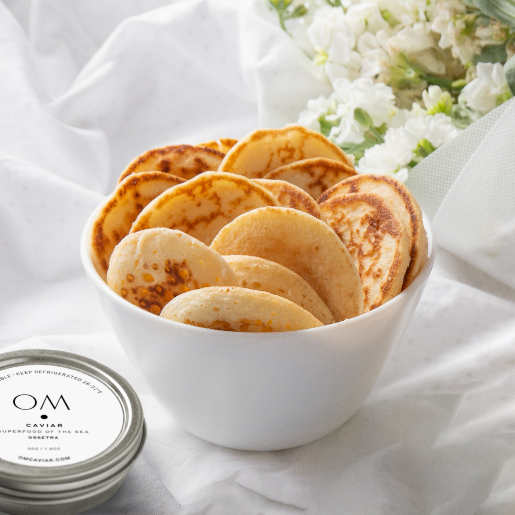 Bowl of golden French mini blinis served with caviar tin, placed on white cloth with white flowers in background
