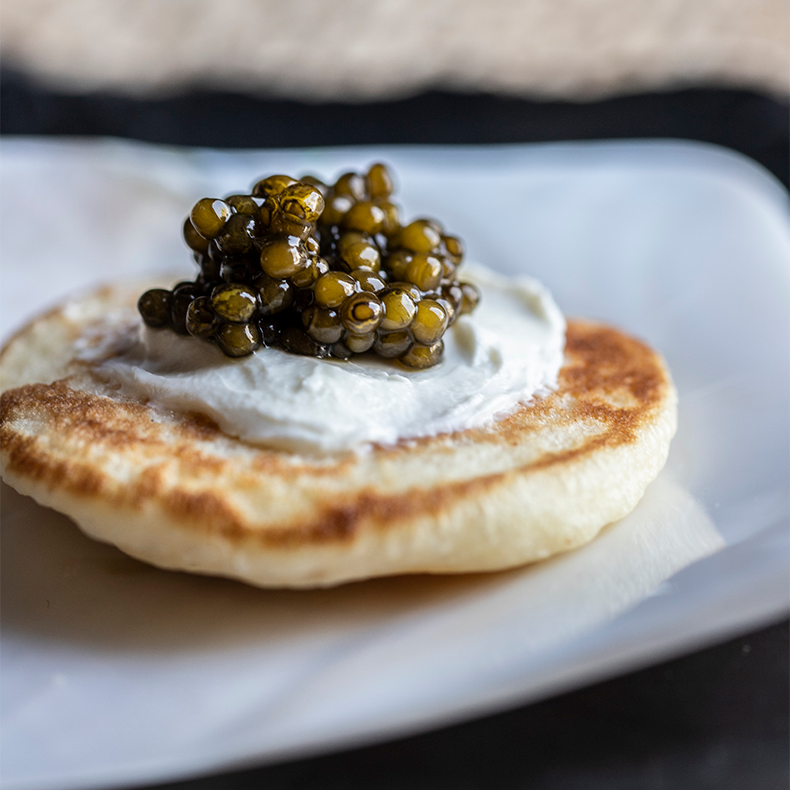 Close-up of a blini topped with cream and Ossetra caviar on a white plate from the Blini Gift Set.